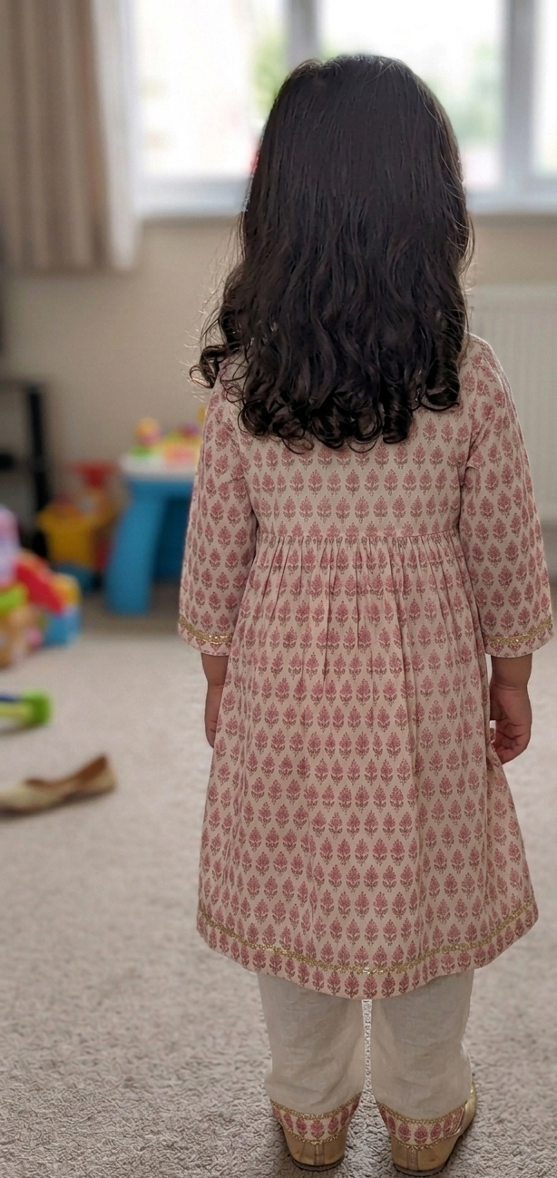 A young child viewed from behind wearing a pink and white patterned tunic dress with cream pants, standing in a carpeted indoor room.