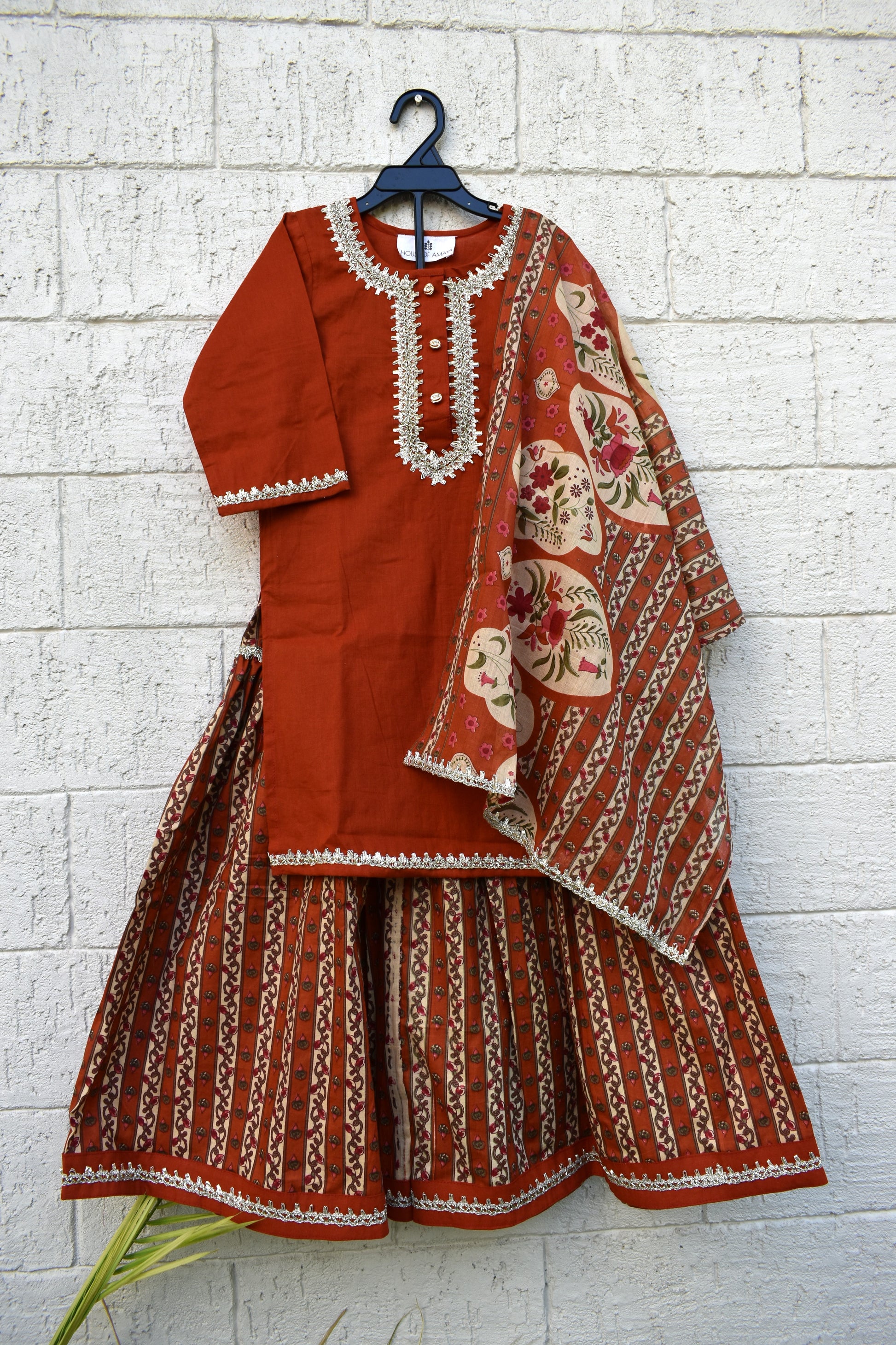 A rust-orange traditional Indian outfit set consisting of a kurta with white embroidery, matching flared pants, and a patterned dupatta, hanging on a black hanger against a white brick wall.