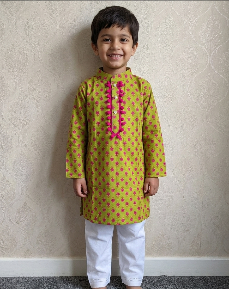 A young boy standing barefoot indoors against a beige patterned wallpaper wall, wearing a yellow kurta with pink floral motifs and a pink decorative placket, paired with white pants.