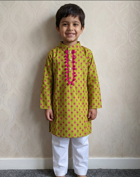 A young boy standing barefoot indoors against a beige patterned wallpaper wall, wearing a yellow kurta with pink floral motifs and a pink decorative placket, paired with white pants.