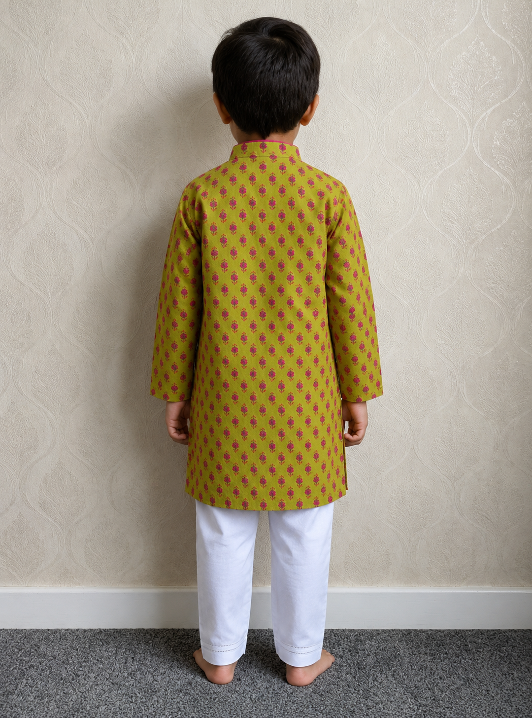 A child standing with their back to the camera, wearing a mustard yellow kurta with a red and green geometric pattern and white pants, set against a beige patterned wallpaper background.