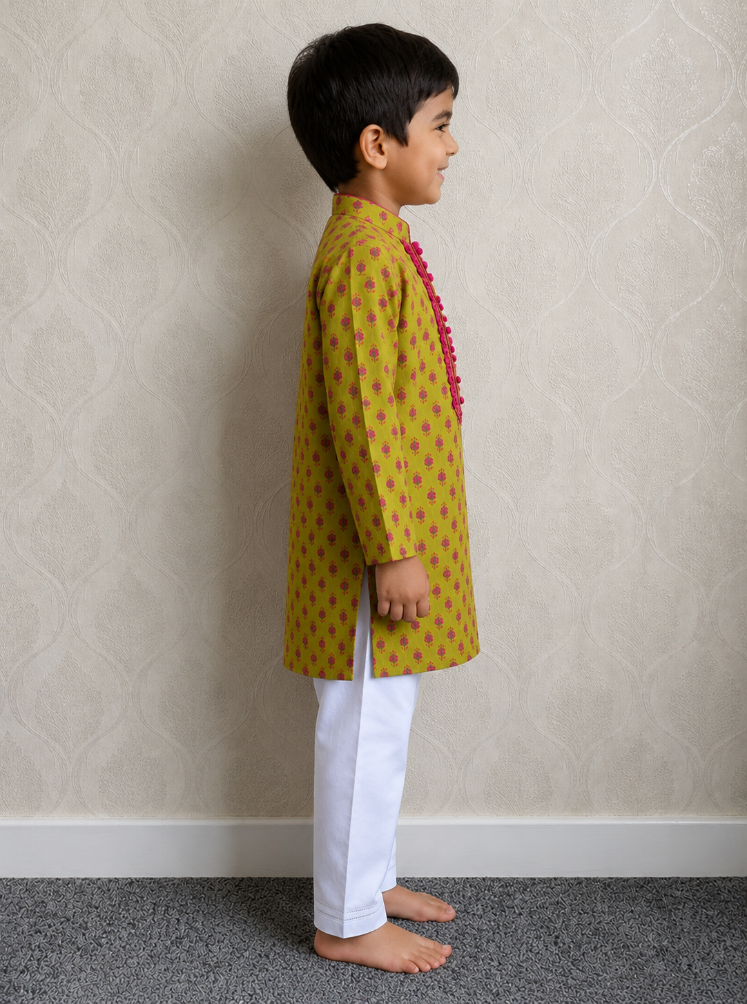 A young boy standing in profile wearing a mustard yellow kurta with small green motifs and a pink tassel detail, paired with white trousers, against a textured beige wall.