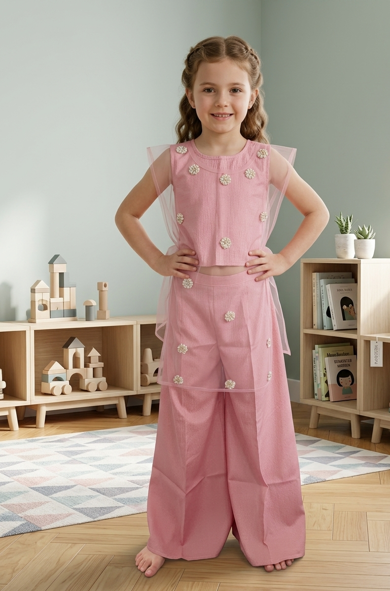 A young girl with braided hair stands barefoot in a child's playroom, wearing a pink sleeveless outfit with sheer mesh panels and decorative white floral embellishments. The outfit features a fitted top with sheer flutter sleeves and matching wide-leg pants.