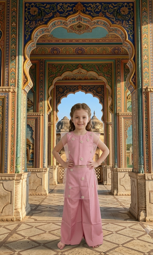 A young girl stands in a grand, ornate architectural corridor wearing a light pink two-piece outfit with a sleeveless top and wide-leg pants. The background features intricate blue, gold, and beige arches.