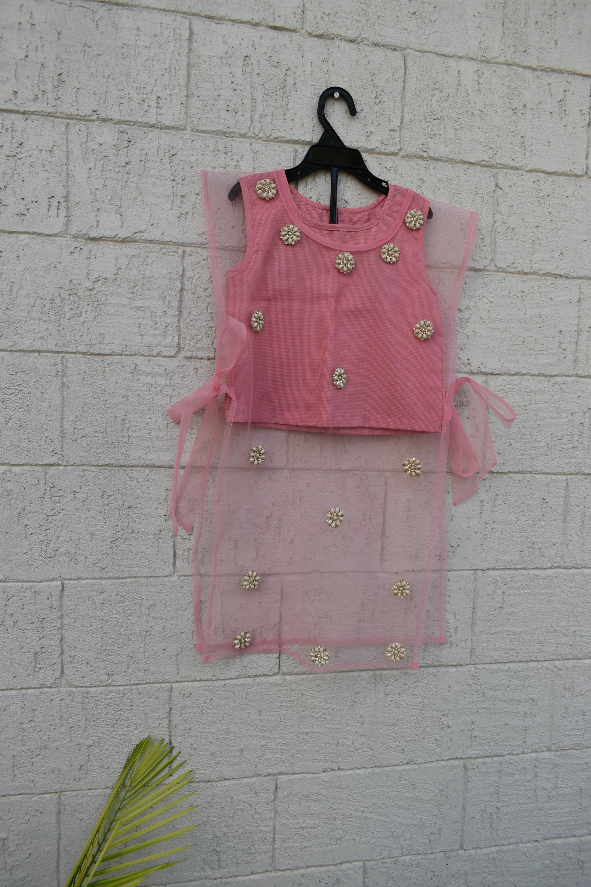 A pink sleeveless baby dress with floral embellishments, displayed on a black hanger against a white painted brick wall.