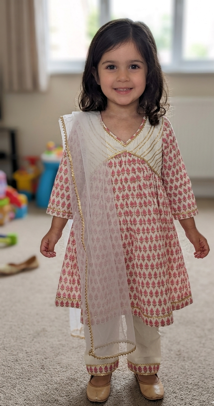 A young girl wearing a traditional Indian salwar kameez outfit with a pink and white floral pattern, paired with a white dupatta featuring gold embroidery, standing in a living room.