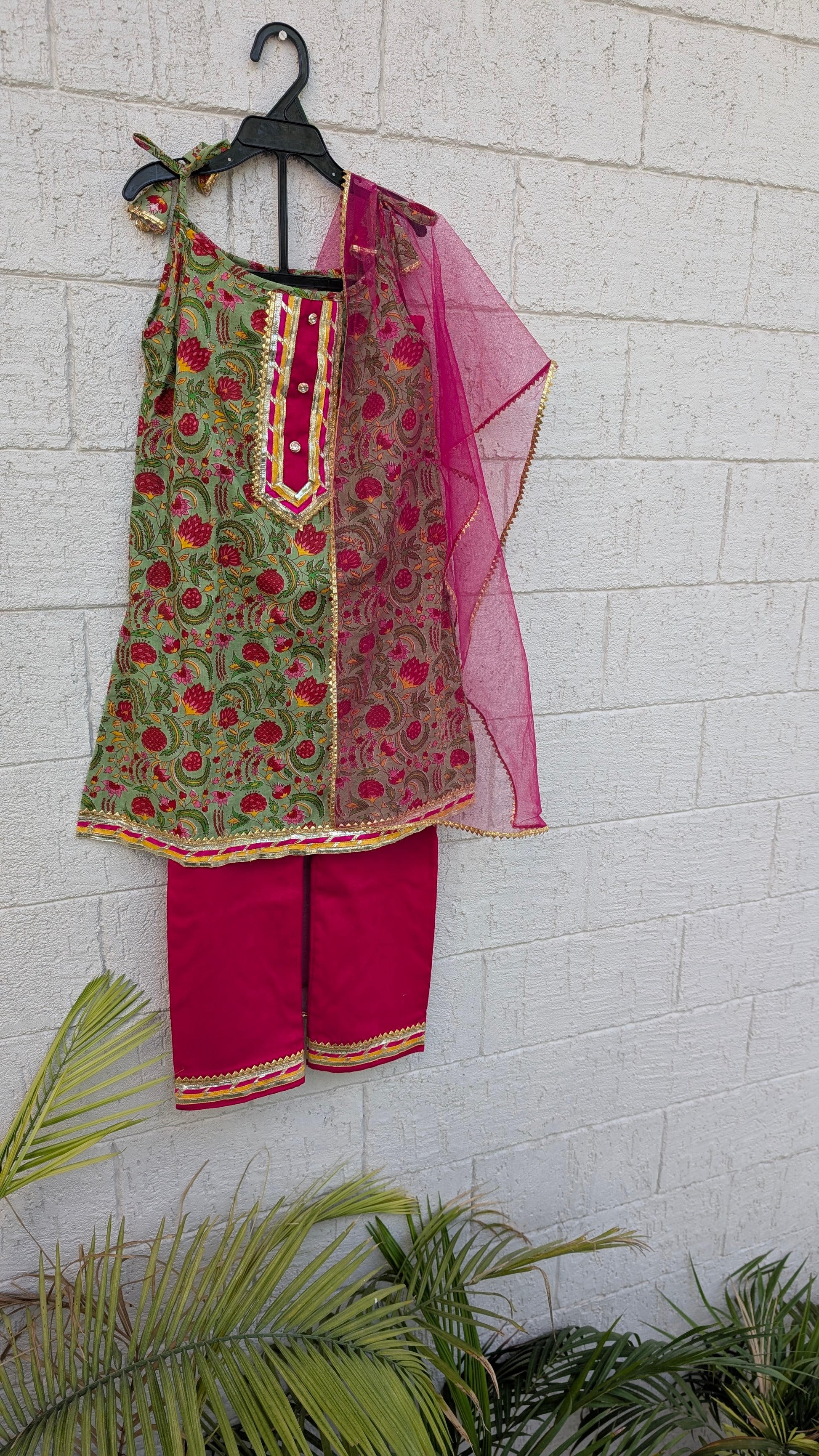 A women's ethnic outfit hanging on a black hanger against a white painted brick wall, featuring a green printed kurta with red floral motifs, bright pink pants, and a sheer pink dupatta.